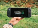 A hand holding a handheld gaming console displaying the Pokémon Legends game screen outdoors.
