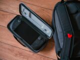 A close-up shot of a gaming console in its case next to a backpack on a wooden floor.