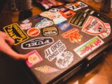 Crop person with leather black suitcase with various stickers placed on wooden table