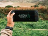 A person holds a handheld gaming device outdoors with Pokémon Legends on screen.