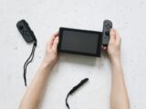 Close-up of hands assembling a Nintendo Switch console with detached controllers on a white surface.