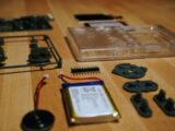 Disassembled electronic components including a clear plastic case, battery, and buttons on wood