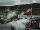 A steamboat with American flags sails through a misty forested lake area.