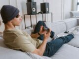 Casual young man playing mobile games on sofa with speakers in the background.